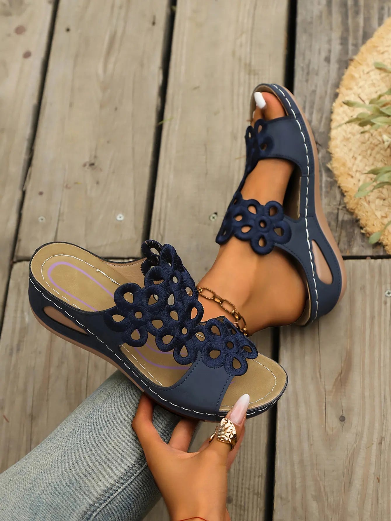Navy blue Zylphra women's wedge sandals with intricate laser-cut floral details on the strap, shown worn on a foot with a straw hat and greenery in the background.