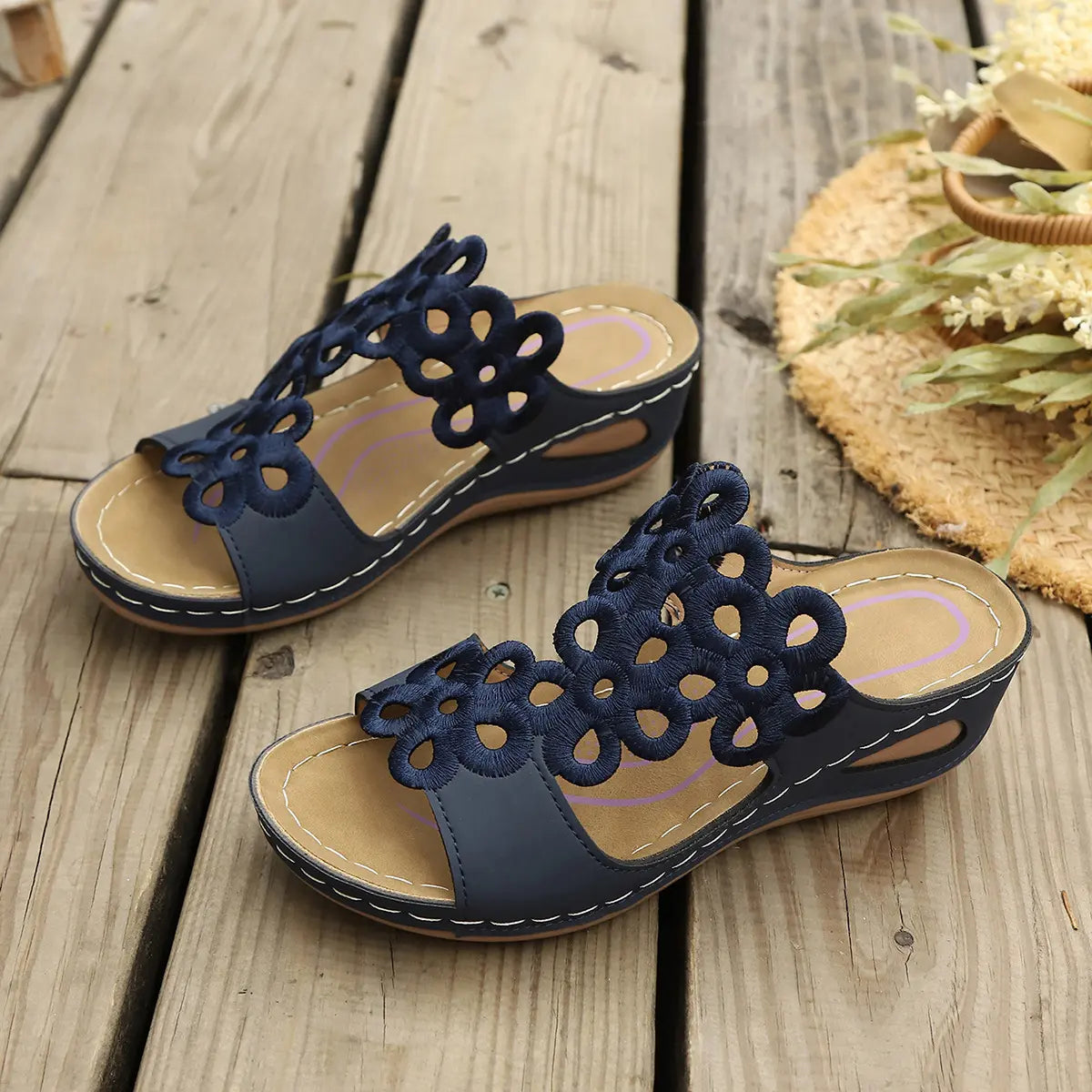 Navy blue Zylphra bohemian wedge sandals for women, featuring intricate laser-cut floral motifs on a wide strap and a comfortable contoured sole, displayed on a rustic wooden plank with a straw hat and dried flowers.