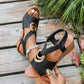 Women's black Zylva wedge sandals with gold ring detail and elastic straps, worn on feet on a wooden deck with lush greenery in the background.