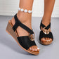 Close-up of women's black Zylva wedge sandals featuring a polished gold-tone ring on elastic straps and a layered wedge heel, shown on feet with white-painted toenails.