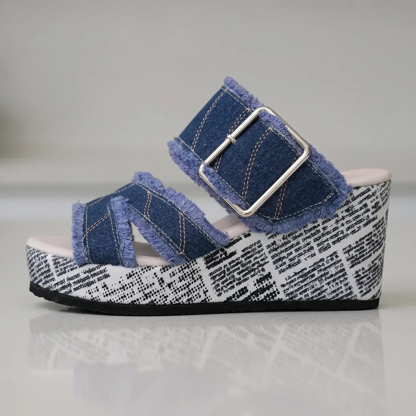 Side view of Zylva women's wedge sandals in blue denim with frayed edges and a silver buckle, featuring a black and white newspaper print on the wedge heel.