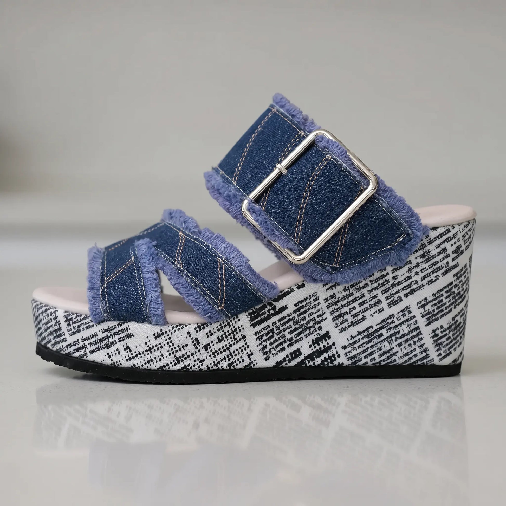 Side view of Zylva women's wedge sandals in blue denim with frayed edges and a silver buckle, featuring a black and white newspaper print on the wedge heel.