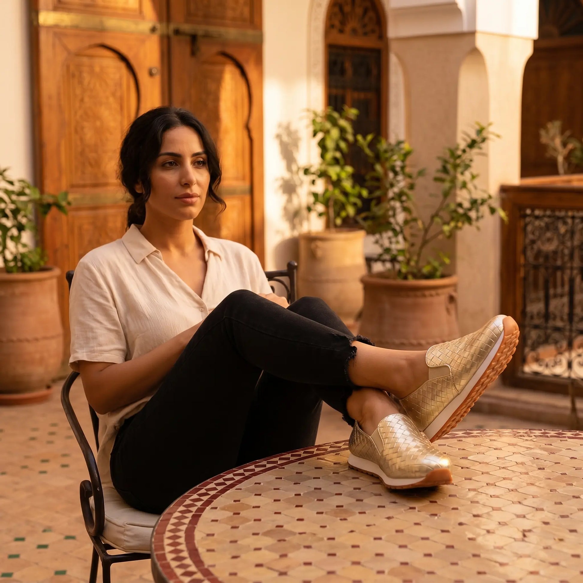 A woman wearing metallic gold woven slip-on loafers with a white and brown rubber sole, seated in a sunlit courtyard with patterned tiles.