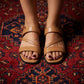 Model's feet showcase Zyneth Desert Tan vegan leather flat sandals, featuring elegant asymmetric crossover straps and an elastic slingback, on a vibrant red patterned rug.