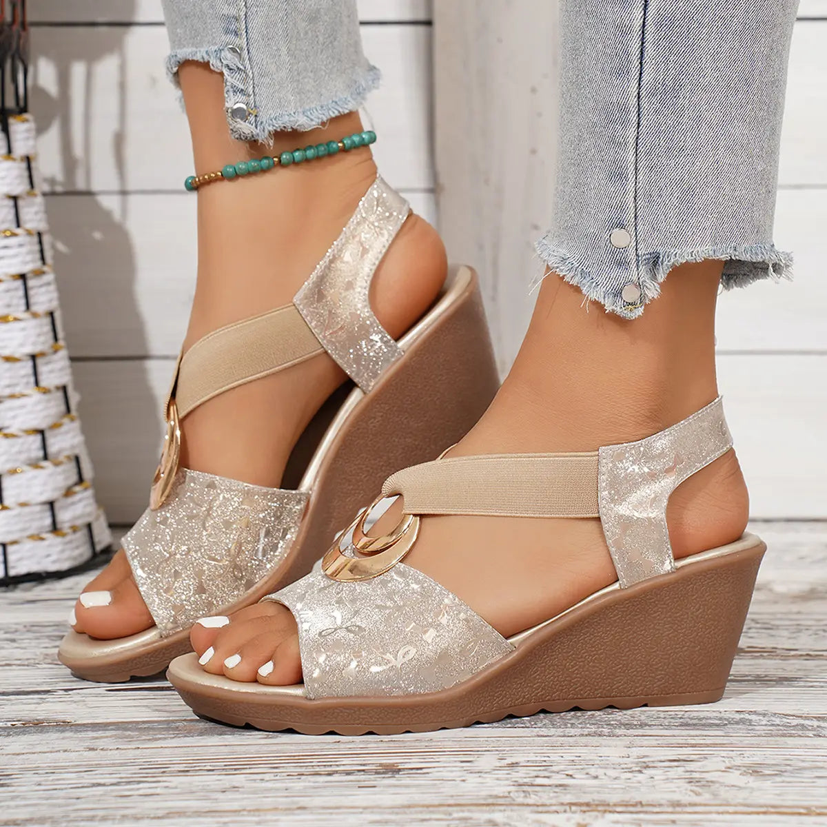 Close-up of shimmering gold floral metallic wedge sandals on a model's feet, featuring elastic criss-cross straps with a gold ring and a peep-toe design with a matte brown wedge heel.