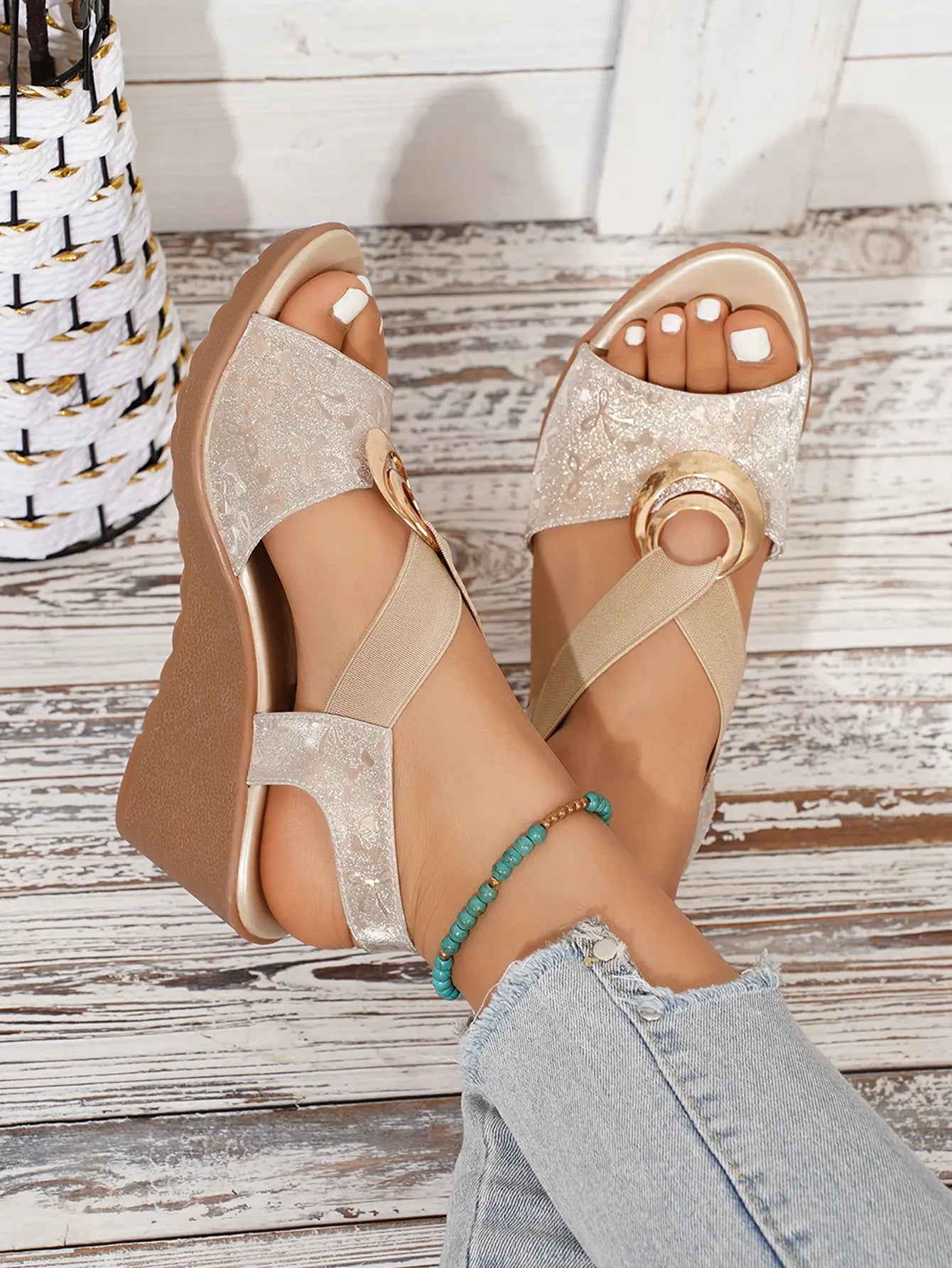 Shimmering metallic floral peep-toe wedge sandals in pale gold worn by a model with white-toe polish and a turquoise anklet, featuring elastic criss-cross straps with a gold-tone ring, set against a white-washed wood plank background.