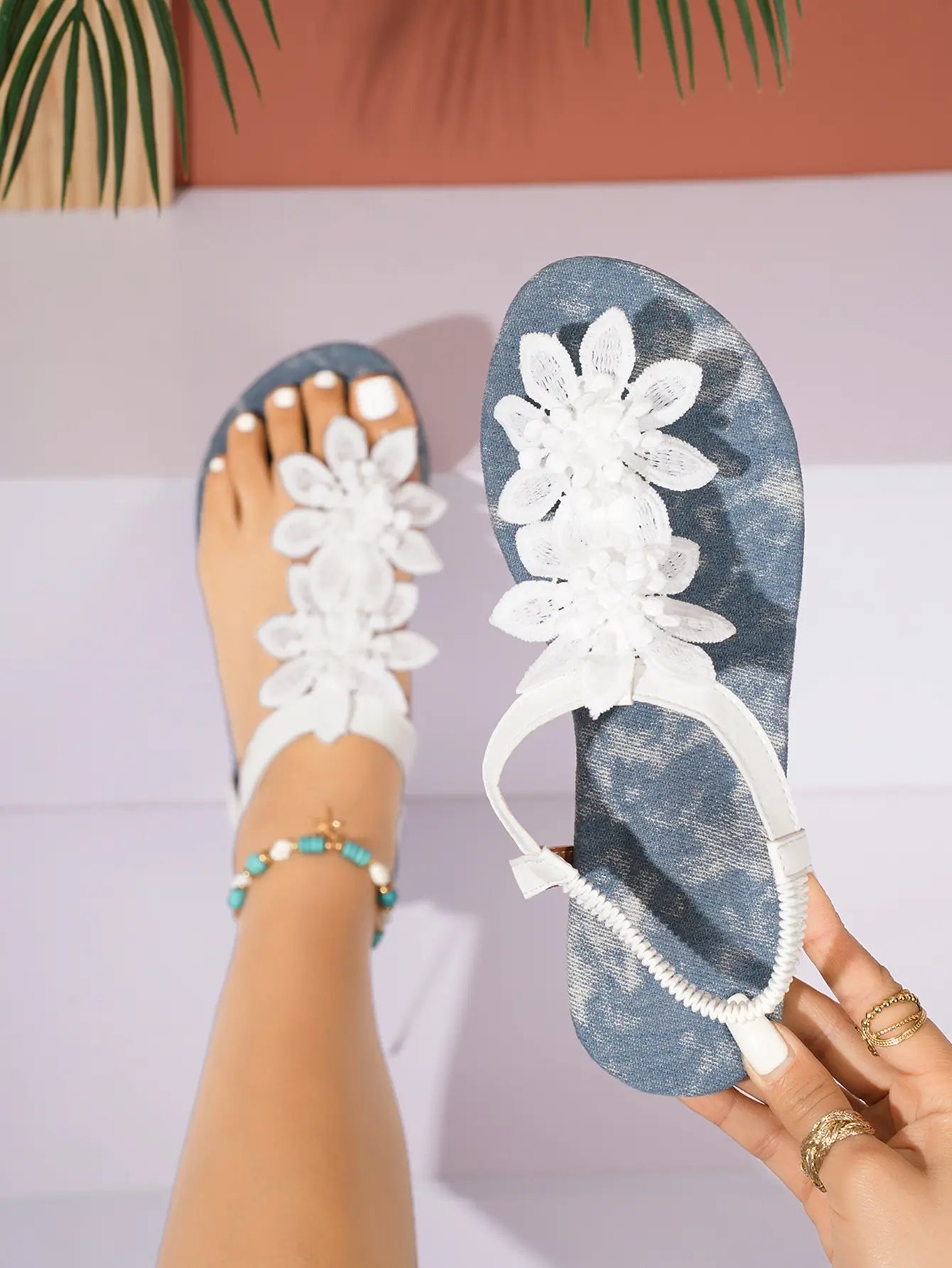 White bohemian thong sandals with delicate floral lace embellishments and elasticated ankle straps, shown on a foot with a turquoise beaded anklet.