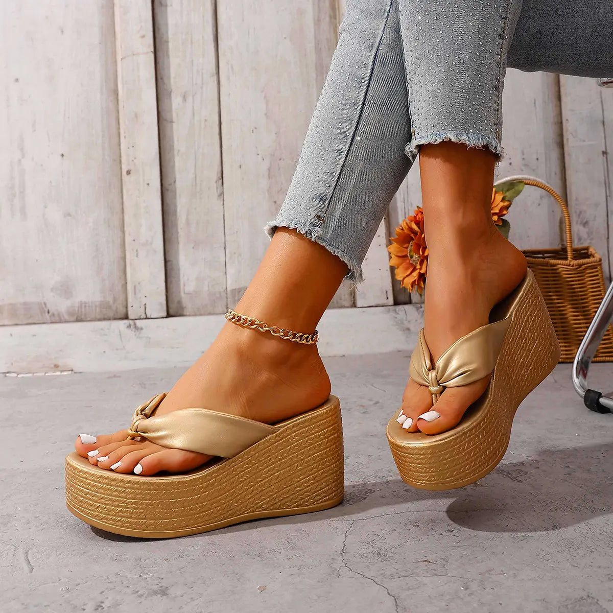 Woman's feet wearing shimmering gold metallic wedge platform flip-flops with a knotted toe strap and woven-effect base, paired with light wash denim jeans.
