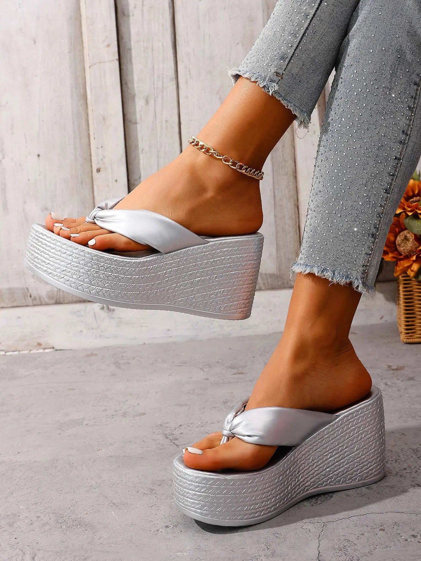 Women's shimmering silver metallic wedge platform flip-flops with a knotted toe strap and woven-textured wedge base, shown on feet with denim jeans.