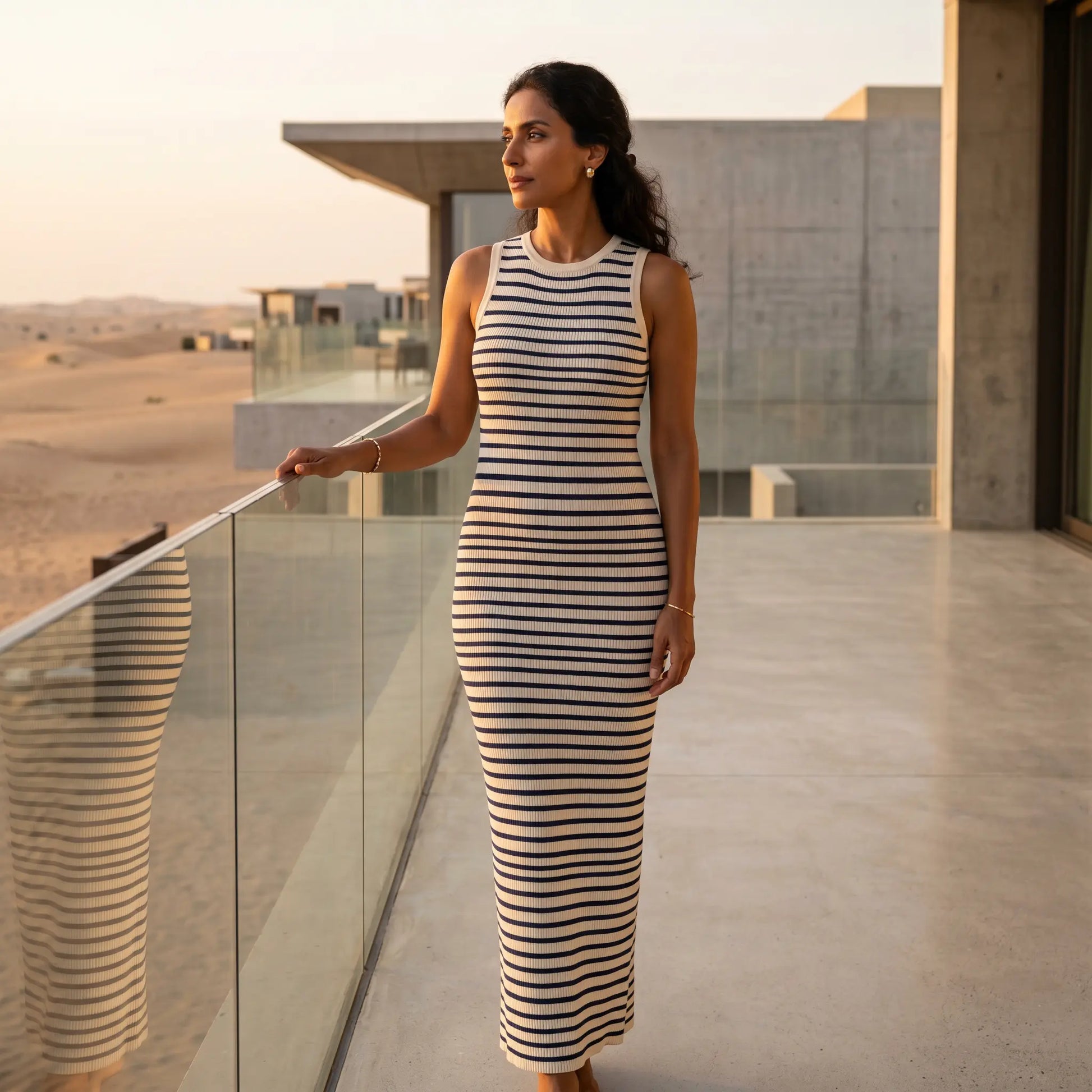 Elegant woman in a Zyphron cream and navy striped ribbed knit maxi dress, showcasing the bodycon fit, on a modern balcony at sunset.