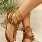 Zyra bohemian deep brown suede-like flat sandals featuring distinctive cross-stitching on the toe strap and braided ankle ties, shown on a model's feet in a lifestyle setting with natural greenery.