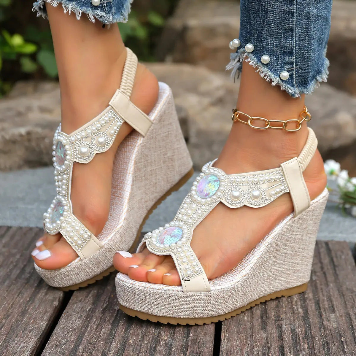 Bohemian off-white embellished wedge sandals featuring faux pearls and iridescent crystals on a T-strap and textured linen-like wedge heel, worn with distressed jeans in an outdoor lifestyle setting.