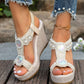Close-up of off-white bohemian wedge sandals adorned with pearls and crystals, worn with embellished denim jeans and a gold anklet on a wooden surface.