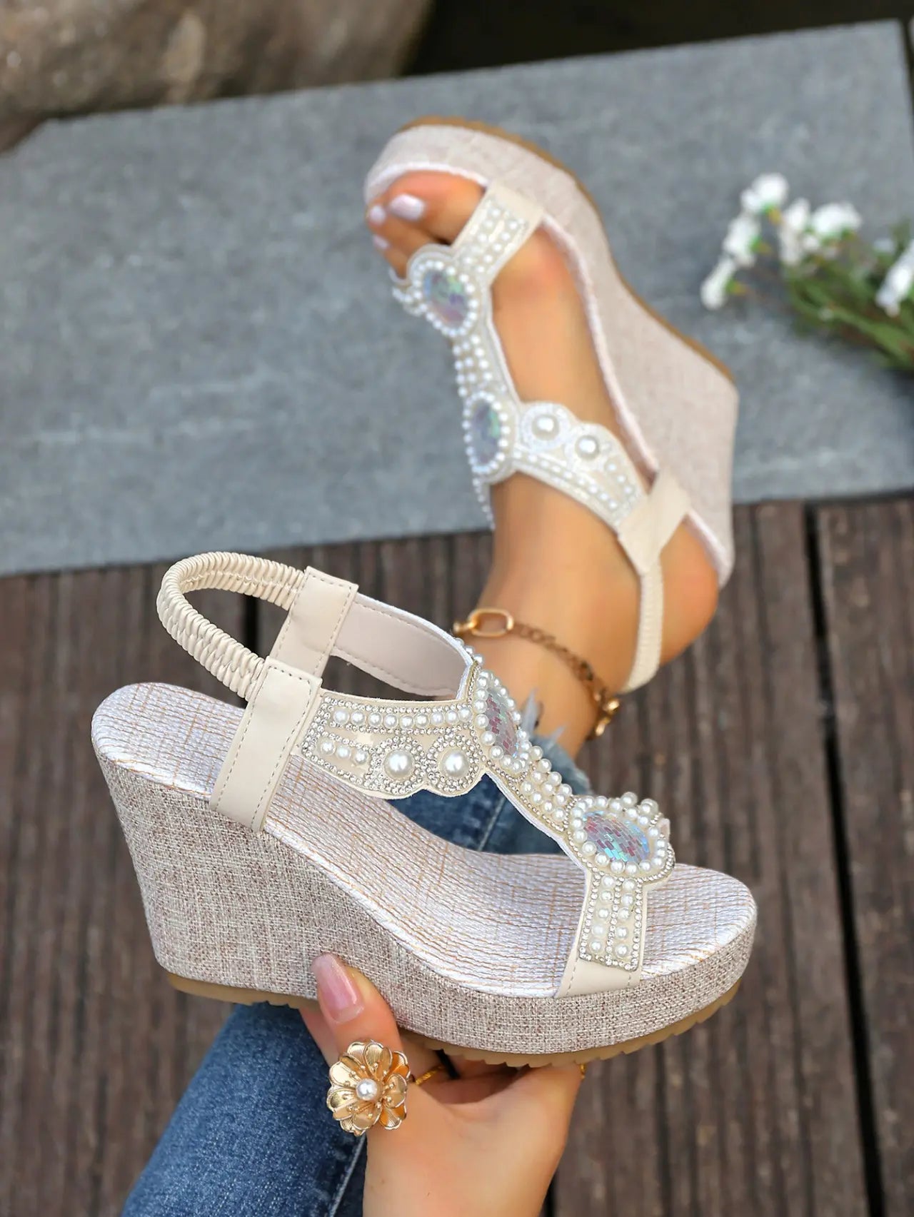 Close-up of an off-white bohemian wedge sandal adorned with faux pearls and iridescent crystals on a T-strap, worn on a foot against a natural wood and stone background. Features a textured woven wedge heel and peep-toe.
