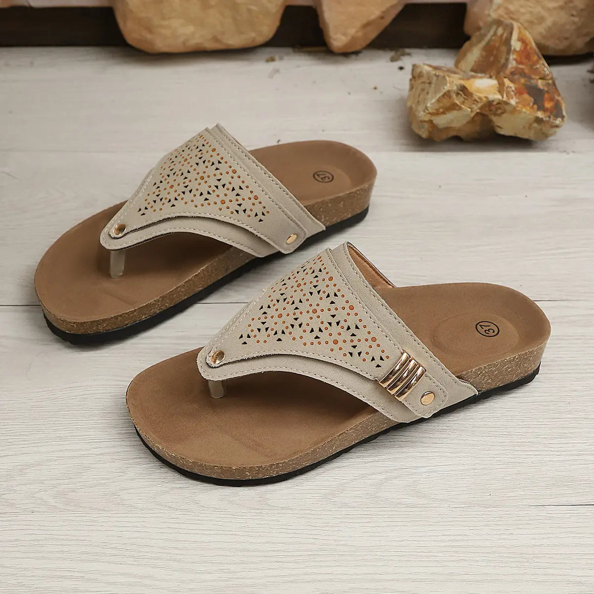 Zyra women's laser-cut thong platform sandals in a neutral taupe color, featuring a cork-patterned footbed and a gold-tone buckle accent. Displayed at a three-quarter angle on a wooden surface with stone decor.