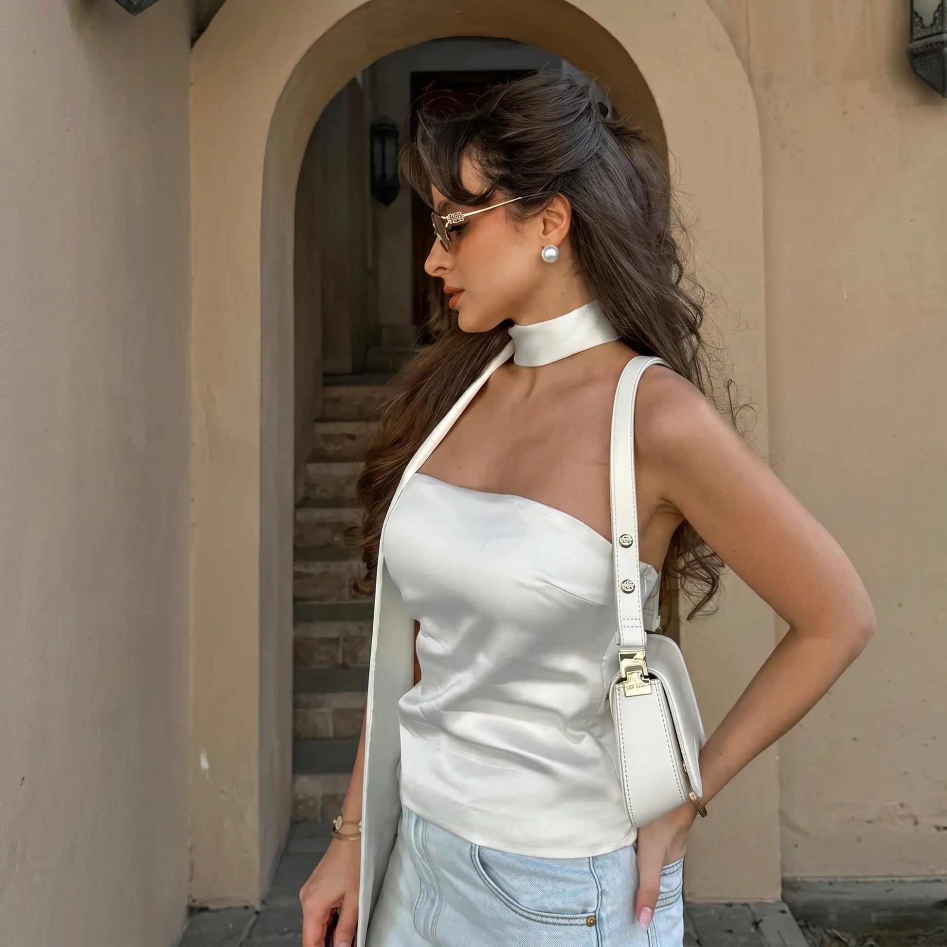 Model wears a white satin bandeau top with integrated halter scarf and light blue denim skirt, styled in an outdoor architectural setting. The top features a subtle sheen and side stud accents.