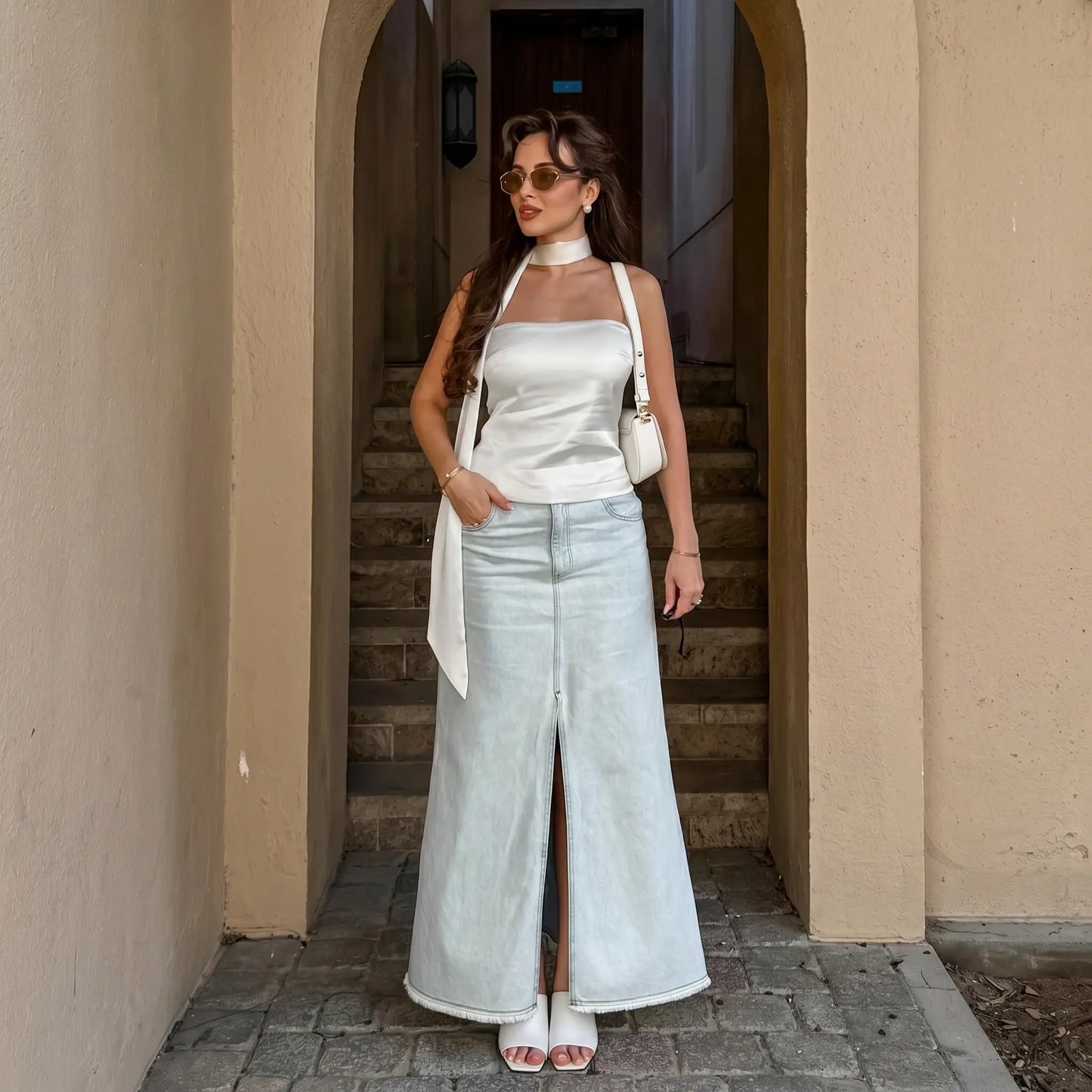 Model wearing a white satin bandeau top with integrated halter scarf and a light wash denim maxi skirt, styled in an outdoor architectural setting. Elegant summer outfit.