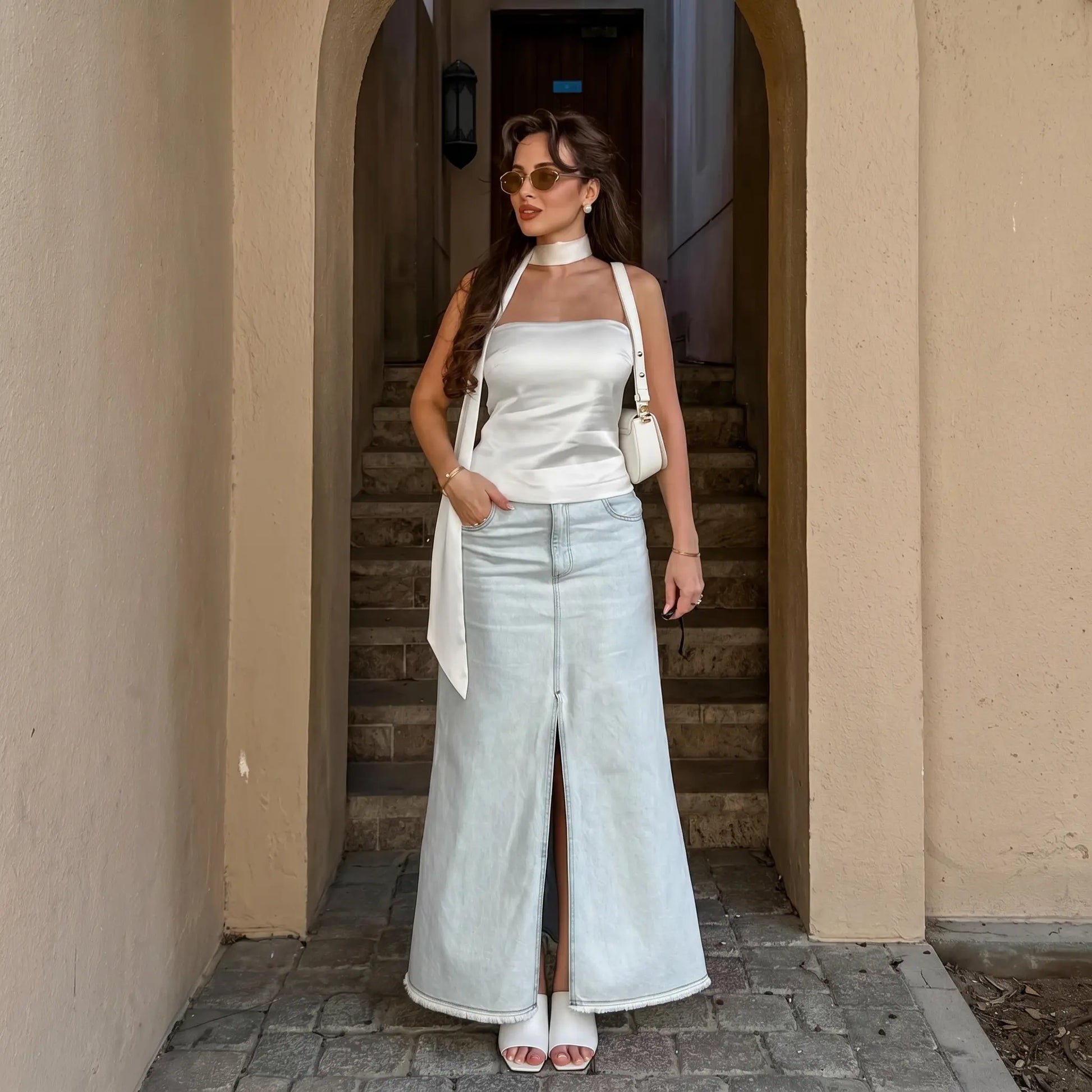 Model wearing a white satin bandeau top with integrated halter scarf and a light wash denim maxi skirt, styled in an outdoor architectural setting. Elegant summer outfit.