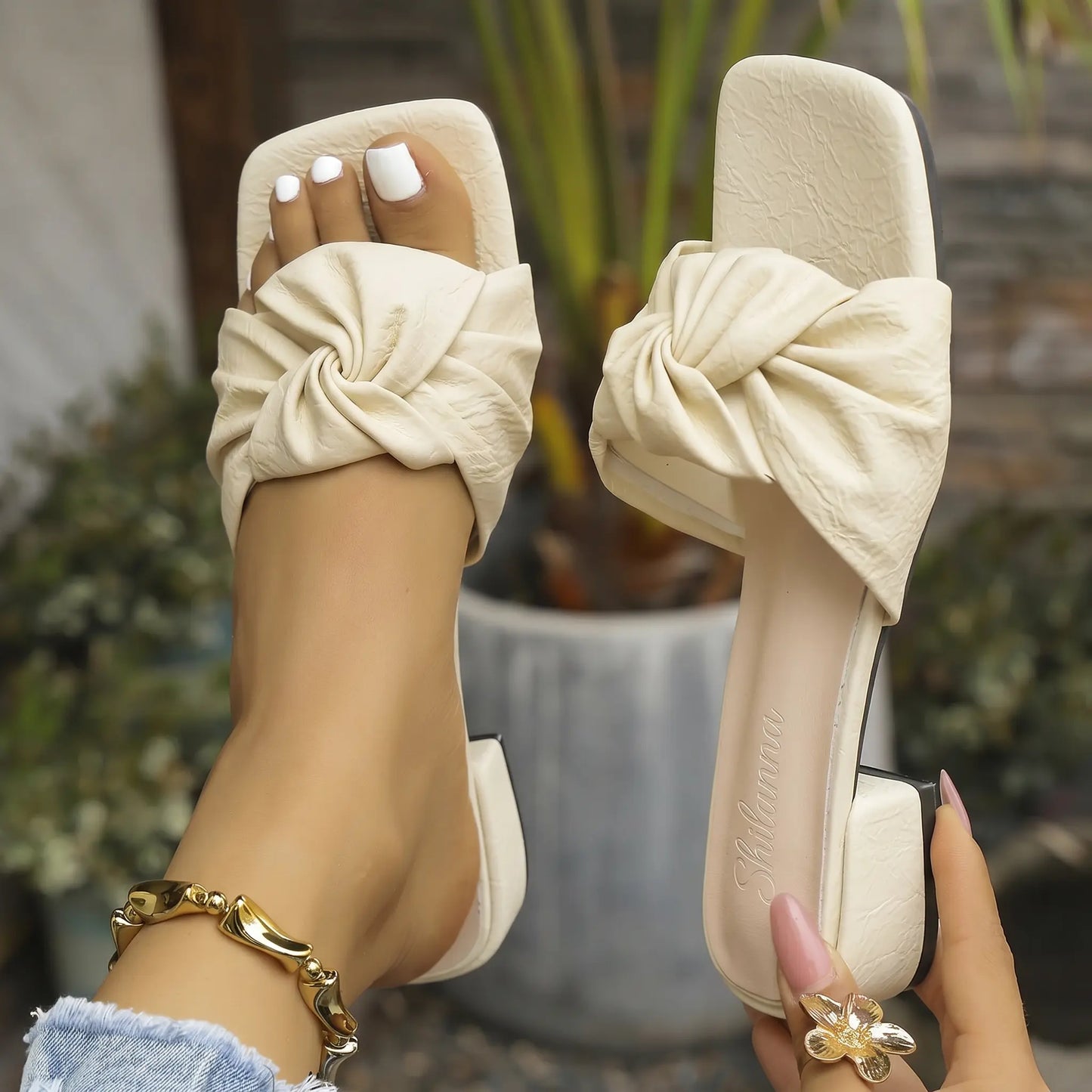 A pair of women's creamy off-white slip-on slide sandals with square toes and a prominent twisted knot detail on the single vamp strap, displayed on a foot with painted white toenails and held by a hand with a gold ring.